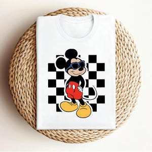 May include: White t-shirt with a black and white checkered pattern and a cartoon image of Mickey Mouse wearing sunglasses. Mickey Mouse is standing with his hands on his hips.