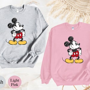 May include: Two sweatshirts with a cartoon drawing of Mickey Mouse. The sweatshirt on the left is ash grey and the sweatshirt on the right is light pink.