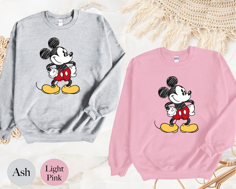 May include: Two sweatshirts with a cartoon drawing of Mickey Mouse. The sweatshirt on the left is ash grey and the sweatshirt on the right is light pink.