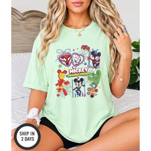 May include: A light green t-shirt featuring a graphic of Mickey Mouse and other characters. The graphic includes the text "Mickey Amazing Friends" and images of Spider-Man, Iron Man, and the Hulk. The shirt is a short-sleeved, casual style.