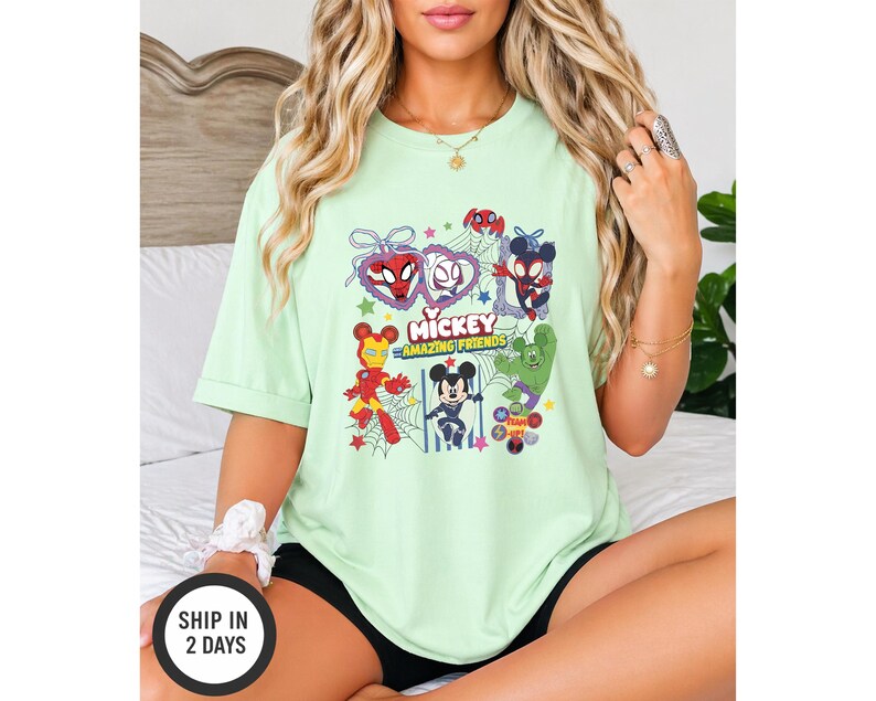 May include: A light green t-shirt featuring a graphic of Mickey Mouse and other characters. The graphic includes the text "Mickey Amazing Friends" and images of Spider-Man, Iron Man, and the Hulk. The shirt is a short-sleeved, casual style.