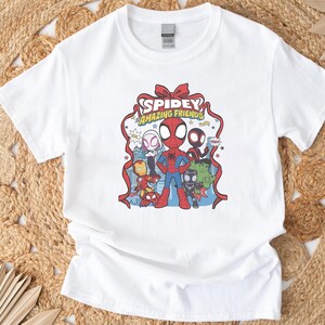 以下が含まれることがあります： スパイディとアメイジング・フレンズのカラフルなグラフィックが特徴の白いTシャツ。デザインにはスパイダーマン、アイアンマン、その他のキャラクターが含まれています。「Spidey Amazing Friends」の文字がキャラクターの上に表示されています。