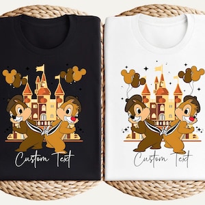 May include: Black and white t-shirts featuring a cartoon illustration of two chipmunks with a castle backdrop. The chipmunks are wearing hats and holding balloons. The text "Custom Text" is written in cursive below the illustration.