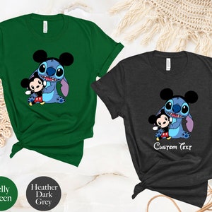 May include: Two t-shirts with a cartoon image of Stitch from Lilo & Stitch holding Mickey Mouse. The shirt on the left is Kelly green and the shirt on the right is heather dark grey. The text "Custom Text" is printed on the grey shirt.