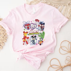 May include: A light pink t-shirt featuring a colorful graphic of Mickey Mouse and various Marvel characters, including Spider-Man, Iron Man, and the Hulk. The text "Mickey Amazing Friends" is also displayed on the shirt.