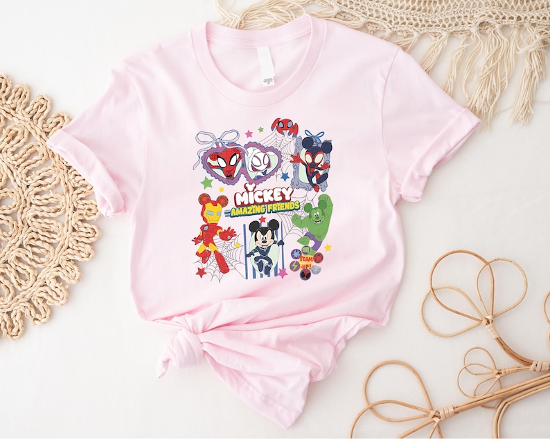 May include: A light pink t-shirt featuring a colorful graphic of Mickey Mouse and various Marvel characters, including Spider-Man, Iron Man, and the Hulk. The text "Mickey Amazing Friends" is also displayed on the shirt.