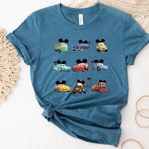 Puede incluir: Una camiseta azul turquesa con un estampado gr&aacute;fico de nueve coches de dibujos animados con orejas de Mickey Mouse. Los coches son rojos, azules, verdes y amarillos.