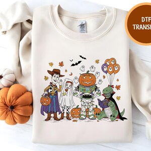 Peut inclure: Sweat-shirt crème avec un motif sur le thème d'Halloween. Le motif comprend des personnages de dessins animés costumés, des citrouilles, des fantômes, des chauves-souris et des ballons. Le texte "DTF TRANSFER" est dans un cercle marron.