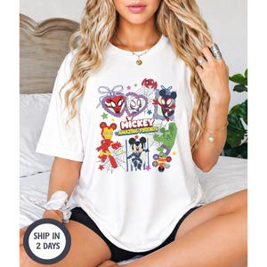 May include: White t-shirt featuring a colorful graphic of Mickey Mouse and Marvel characters, including Spider-Man, Iron Man, and the Hulk. The text "Mickey Amazing Friends" is also displayed. The shirt has short sleeves.