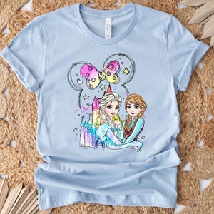 May include: Light blue t-shirt with a colorful illustration of Elsa and Anna from Frozen, standing in front of a castle with a pink bow and stars.