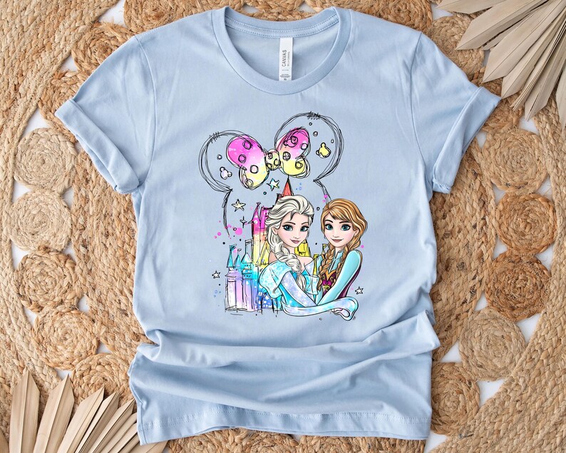 May include: Light blue t-shirt with a colorful illustration of Elsa and Anna from Frozen, standing in front of a castle with a pink bow and stars.