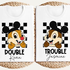 May include: Two white t-shirts with a cartoon chipmunk design. Each shirt features a different name below the chipmunk, with the words "DOUBLE TROUBLE" above. The chipmunk has a red nose and is wearing a Mickey Mouse hat.