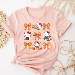 Può includere: Una maglietta rosa chiaro con un design grafico di personaggi di Hello Kitty e fiocchi a tema autunnale. Il design include Hello Kitty con un tacchino, che tiene un cestino e circondata da zucche e foglie.