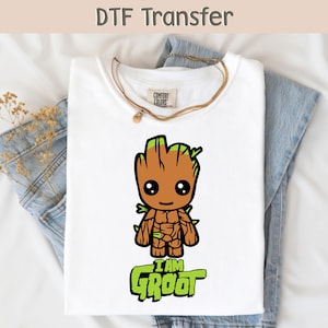 以下が含まれることがあります： 白いTシャツ。漫画のグルートのイラストと緑色の「I AM GROOT」の文字が描かれています。柔らかい素材で作られており、白い表面に青いジーンズが添えられています。