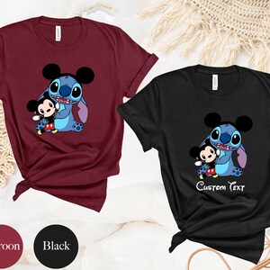 May include: Two t-shirts, one maroon and one black, featuring a cartoon image of Stitch from Lilo & Stitch holding a Mickey Mouse character. The black t-shirt has the text "Custom Text" printed below the image.
