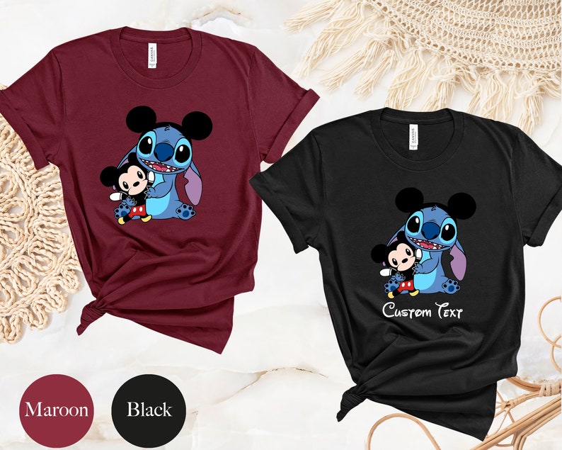 May include: Two t-shirts, one maroon and one black, featuring a cartoon image of Stitch from Lilo & Stitch holding a Mickey Mouse character. The black t-shirt has the text "Custom Text" printed below the image.