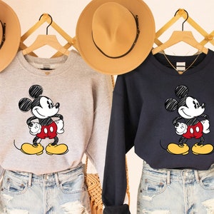 May include: Two grey and navy blue sweatshirts with a cartoon drawing of Mickey Mouse in black and white. Mickey Mouse is wearing red shorts and yellow shoes. The sweatshirts are on hangers and have a V-neck.
