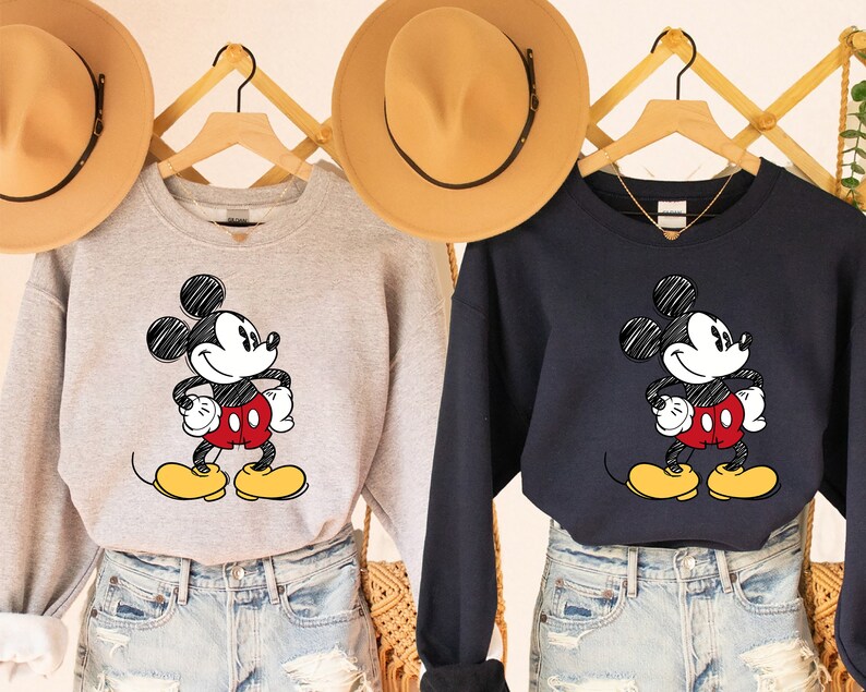 May include: Two grey and navy blue sweatshirts with a cartoon drawing of Mickey Mouse in black and white. Mickey Mouse is wearing red shorts and yellow shoes. The sweatshirts are on hangers and have a V-neck.
