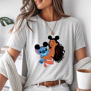 May include: Light gray t-shirt featuring a graphic of a cartoon character hugging a blue alien. The cartoon character has black hair and is wearing a red top. The alien is blue with large eyes. The shirt has a crew neck and short sleeves.