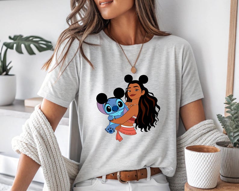 May include: Light gray t-shirt featuring a graphic of a cartoon character hugging a blue alien. The cartoon character has black hair and is wearing a red top. The alien is blue with large eyes. The shirt has a crew neck and short sleeves.