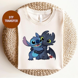May include: A cream-colored t-shirt featuring a cartoon graphic of Stitch and Toothless hugging. Stitch is blue with large ears, and Toothless is black with green eyes. The shirt has a round neck and short sleeves. A brown circle with the text "DTF TRANSFER" is in the upper left corner.