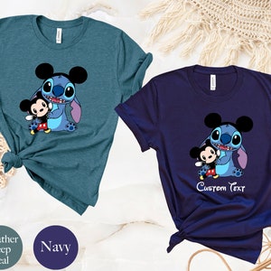 May include: Two t-shirts featuring a cartoon image of Stitch from Lilo & Stitch hugging Mickey Mouse. The shirt on the left is a heather deep teal color. The shirt on the right is navy blue and has the text "Custom Text" printed below the image.