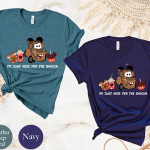 May include: Two t-shirts with a cartoon image of a tow truck with Mickey Mouse ears, holding a cup of coffee, a donut, and a Mickey Mouse apple. The text on the shirts reads "I'm just here for the snacks". The shirts are available in heather deep teal and navy.