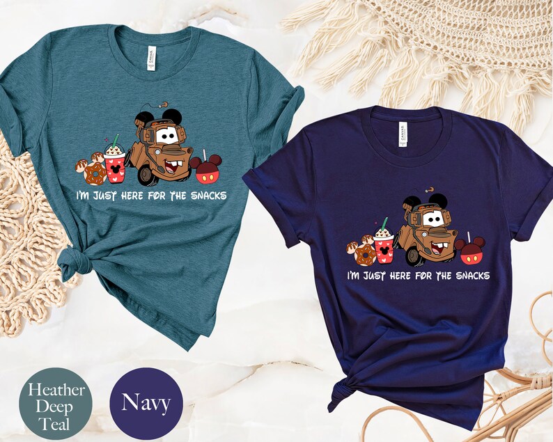 May include: Two t-shirts with a cartoon image of a tow truck with Mickey Mouse ears, holding a cup of coffee, a donut, and a Mickey Mouse apple. The text on the shirts reads "I'm just here for the snacks". The shirts are available in heather deep teal and navy.