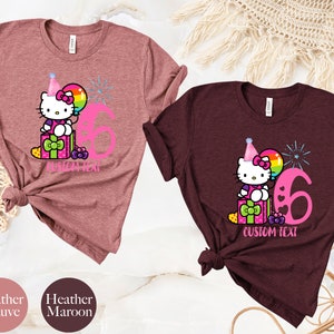 May include: Two t-shirts with a Hello Kitty design. The shirt on the left is heather mauve and the shirt on the right is heather maroon. Both shirts feature Hello Kitty wearing a party hat and holding a balloon. The number 6 is printed on both shirts. The text "CUSTOM TEXT" is printed on both shirts.