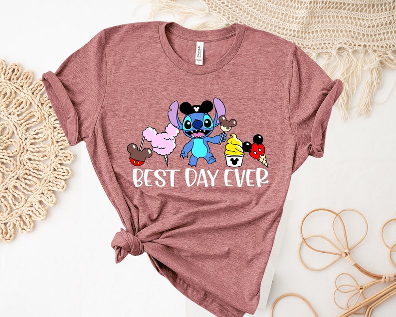 May include: A maroon t-shirt with a cartoon image of Stitch from Lilo & Stitch wearing Mickey Mouse ears and holding a Mickey Mouse ice cream cone. The text "BEST DAY EVER" is printed below the image.