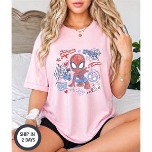 Puede incluir: Una camiseta rosa claro con un dise&ntilde;o de dibujos animados de Spider-Man. El gr&aacute;fico incluye las palabras "Amazing" y "Spider" junto con otros garabatos. La camiseta es de manga corta y de estilo casual.