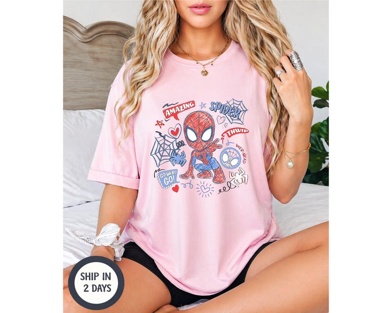 Puede incluir: Una camiseta rosa claro con un dise&ntilde;o de dibujos animados de Spider-Man. El gr&aacute;fico incluye las palabras "Amazing" y "Spider" junto con otros garabatos. La camiseta es de manga corta y de estilo casual.