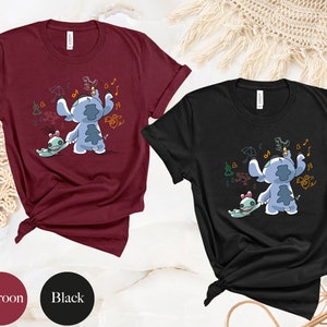 May include: Two t-shirts featuring a blue Stitch character from Lilo & Stitch. Stitch is holding a small green alien and drawing on a wall. The t-shirts are available in maroon and black.