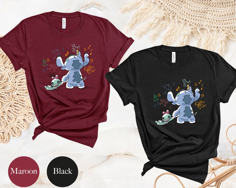 May include: Two t-shirts featuring a blue Stitch character from Lilo & Stitch. Stitch is holding a small green alien and drawing on a wall. The t-shirts are available in maroon and black.