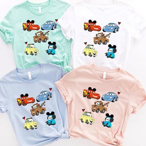 May include: Four different colored t-shirts with a Cars and Mickey Mouse theme. The shirts are light blue, light green, white, and pink. Each shirt features a different combination of Cars characters, including Lightning McQueen, Mater, and Tow Mater. Mickey Mouse is featured on each shirt with a different colored hat.