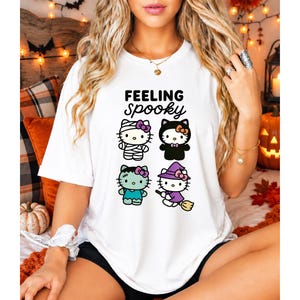 May include: A white t-shirt featuring the text "FEELING SPOOKY" in black lettering above four cartoon cat characters dressed in Halloween costumes: a mummy, a black cat, a Frankenstein monster, and a witch riding a broomstick.
