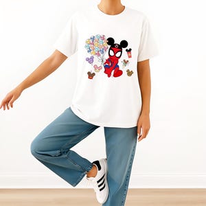 Puede incluir: Camiseta blanca con un personaje de dibujos animados que combina a Spider-Man y Mickey Mouse, sosteniendo globos de colores. La camiseta se combina con vaqueros azules y zapatillas blancas.