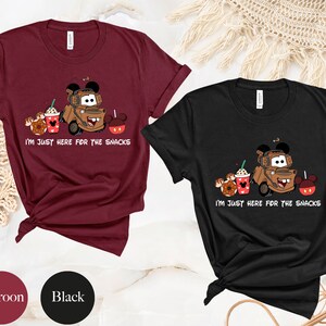 May include: Two t-shirts, one maroon and one black, with a cartoon image of Mater from Cars with Mickey Mouse ears, holding a coffee cup and a donut. The text on the shirts reads "I'm just here for the snacks".