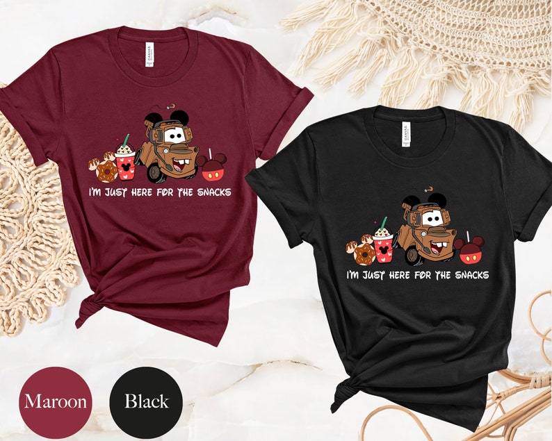 May include: Two t-shirts, one maroon and one black, with a cartoon image of Mater from Cars with Mickey Mouse ears, holding a coffee cup and a donut. The text on the shirts reads "I'm just here for the snacks".