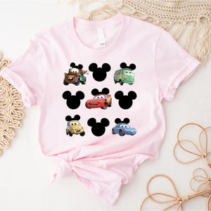 Pu&ograve; includere: Maglietta rosa con orecchie di Topolino nere e immagini di auto del film Disney Cars. Le auto includono Mater, Saetta McQueen e un'auto blu.