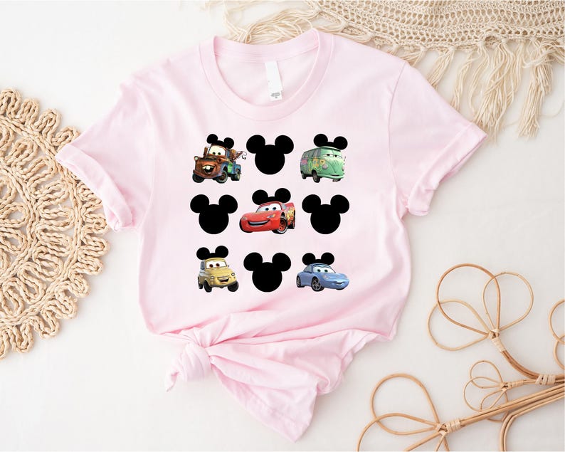 Pu&ograve; includere: Maglietta rosa con orecchie di Topolino nere e immagini di auto del film Disney Cars. Le auto includono Mater, Saetta McQueen e un'auto blu.
