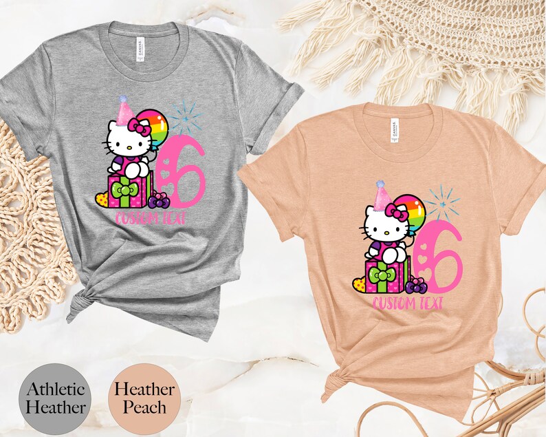 May include: Gray and peach t-shirts with a pink Hello Kitty character wearing a party hat and holding a rainbow balloon. The shirts have a pink number 6 and the text "CUSTOM TEXT".