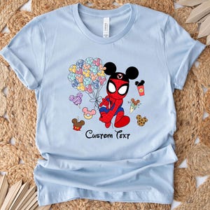 Puede incluir: Camiseta azul claro con un personaje de dibujos animados que combina a Spider-Man y Mickey Mouse, sosteniendo globos de colores. La camiseta tiene la frase "Custom Text" impresa, junto con comida de dibujos animados.