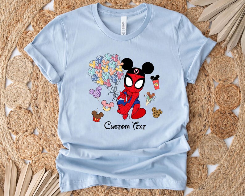Puede incluir: Camiseta azul claro con un personaje de dibujos animados que combina a Spider-Man y Mickey Mouse, sosteniendo globos de colores. La camiseta tiene la frase "Custom Text" impresa, junto con comida de dibujos animados.