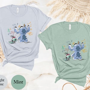May include: Two t-shirts featuring a blue cartoon character with a green friend. The character is holding a pencil and drawing musical notes, raindrops, and an umbrella. The t-shirts are in light blue and mint green.