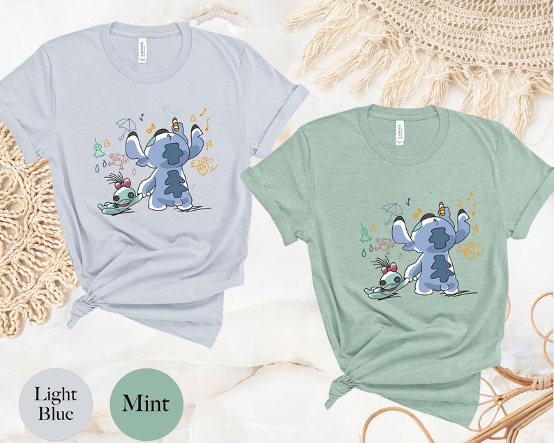 May include: Two t-shirts featuring a blue cartoon character with a green friend. The character is holding a pencil and drawing musical notes, raindrops, and an umbrella. The t-shirts are in light blue and mint green.
