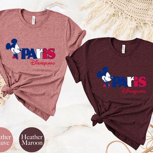 Puede incluir: Dos camisetas con un gr&aacute;fico de Mickey Mouse y el texto "Paris Disneyland". La camiseta de la izquierda es de color malva jaspeado y la camiseta de la derecha es de color marr&oacute;n jaspeado.
