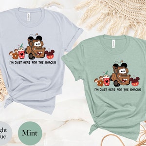 May include: Two t-shirts with a cartoon image of Mater from Cars, a donut, a Mickey Mouse ear ice cream cup, and a Mickey Mouse ear ice cream bar. The text on the shirts reads "I'm just here for the snacks". The shirts are in light blue and mint green.
