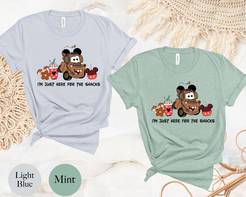 May include: Two t-shirts with a cartoon image of Mater from Cars, a donut, a Mickey Mouse ear ice cream cup, and a Mickey Mouse ear ice cream bar. The text on the shirts reads "I'm just here for the snacks". The shirts are in light blue and mint green.