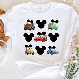 Pu&ograve; includere: Maglietta bianca con una grafica in bianco e nero di orecchie di Topolino e personaggi di Cars. I personaggi di Cars includono Mater, Saetta McQueen e un'auto da corsa blu.
